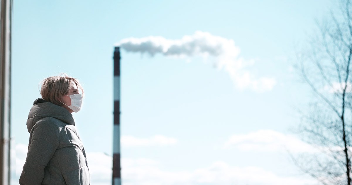 Woman in a face mask standing near a smoking factory chimney